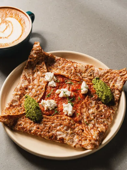 A savory crepe with tomato sauce, pesto, and feta cheese, paired with a latte. This image highlights a cafe's savory lunch options alongside its coffee menu.