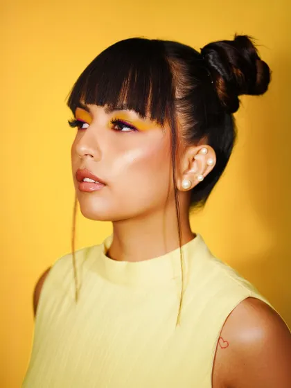 A beautiful profile shot from the "Summer Edit." The look features a high bun, trendy bangs, and a pop of yellow eyeshadow, underlined with a hint of purple for a creative twist.