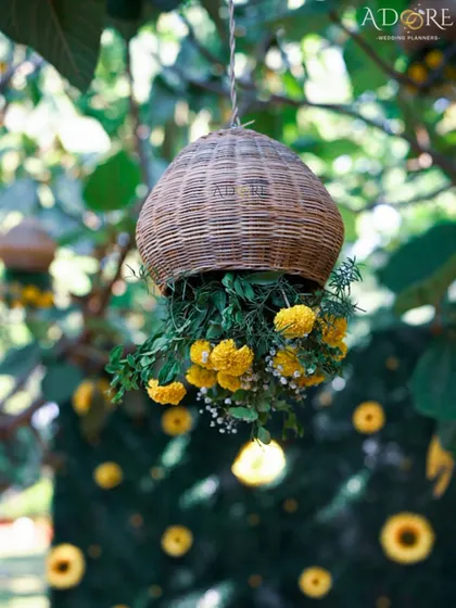 A charming decor detail for an outdoor Haldi. A rustic wicker basket filled with marigolds hangs from a tree, adding to the natural and festive ambiance of the sunflower-themed celebration.