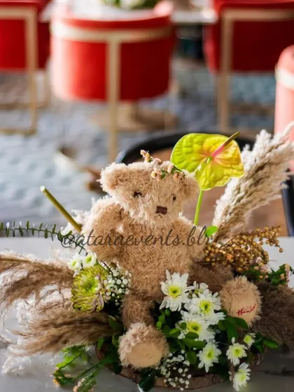 A teddy bear centerpiece with pampas grass and white flowers. This arrangement, set against the backdrop of modern red chairs, shows how I can blend themes with any venue.