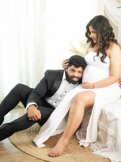 A tender moment between partners in a bright, airy studio setting. His gentle listening to the baby bump is a classic expression of love and anticipation.