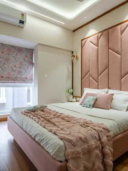 A cozy bedroom with a large, geometrically patterned pink headboard that serves as a statement piece. The floral Roman blinds and soft pink bedding create a romantic and inviting atmosphere.