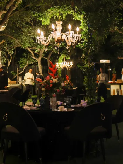 An outdoor seating area for a cocktail night, where tables are set under trees adorned with hanging chandeliers, blending nature with pure opulence.