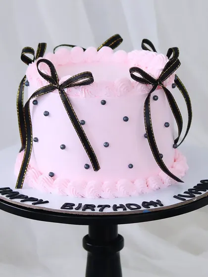 A trendy Coquette-style cake, featuring soft pink buttercream, black polka dots, and delicate black ribbons tied into bows. This design is perfect for a chic and feminine celebration.