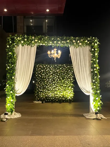 A stunning floral archway at the entrance. The structure is covered in white flowers and greenery, with drapes that can be opened to reveal a beautiful photo wall.