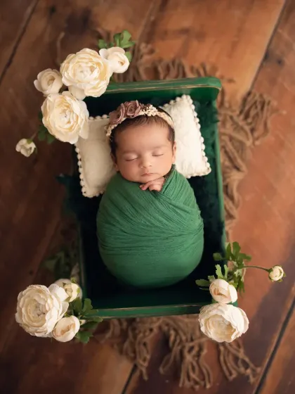 This newborn girl is swaddled in a beautiful emerald green wrap, lying in a matching green box and surrounded by white roses.