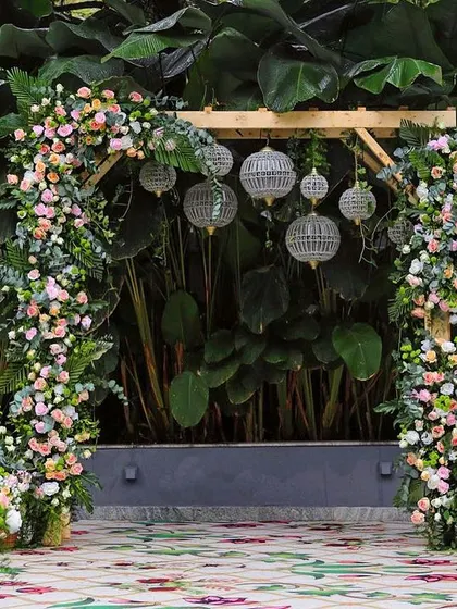 A beautiful floral arch against a backdrop of giant green leaves. The pastel-colored flowers and hanging lanterns create a soft and romantic photo spot.
