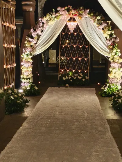 We designed this elegant entrance for a Roka ceremony. The walkway features a white carpet, a floral arch with pastel flowers and drapes, and candle-lit stands, creating a romantic and welcoming path.