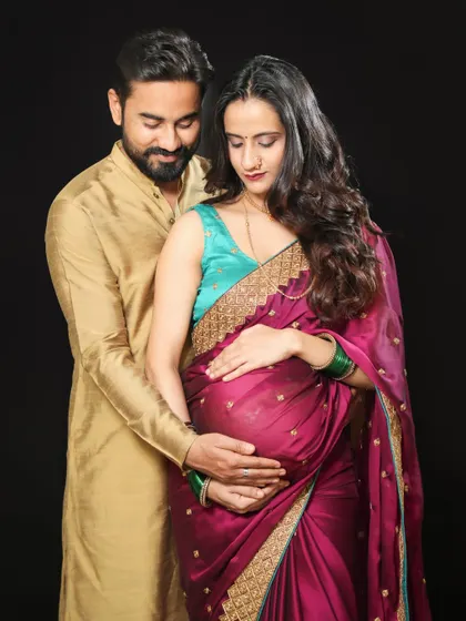 A tender moment where both parents-to-be cradle the baby bump. Dressed in traditional attire against a simple black background, their love and anticipation are the main focus.