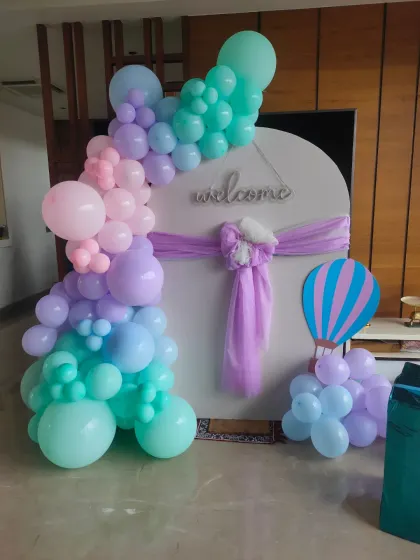 A modern and elegant naming ceremony decoration with a pastel balloon garland on a white circular backdrop. A small hot air balloon prop adds a whimsical touch.