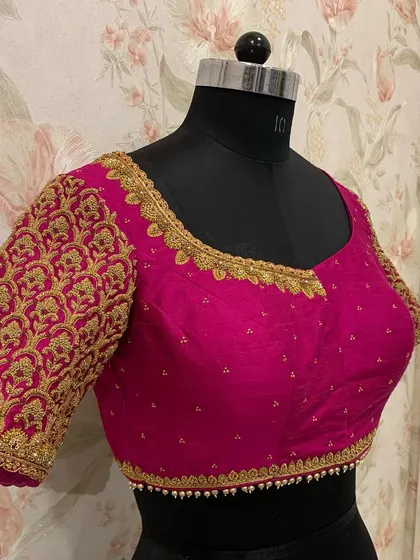 A beautiful magenta bridal blouse with dense, traditional gold embroidery covering the entire sleeve. The small pearl detailing at the hem adds a classic finish.