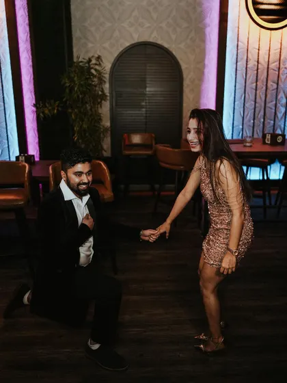 A playful moment where the groom is on one knee, and the bride is laughing and pulling him up, captured at their cocktail party.