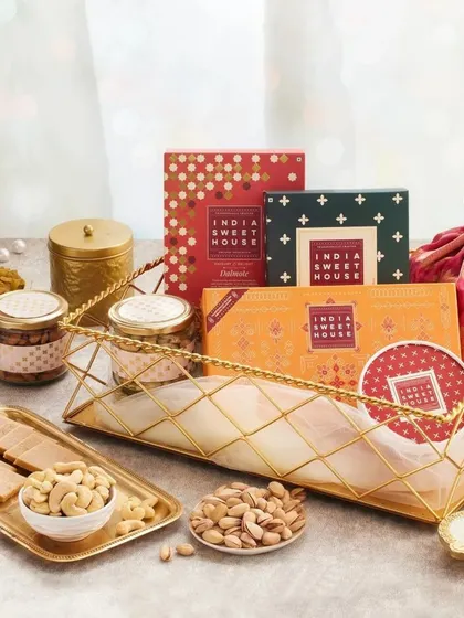 An elegant festive tray featuring boxes from the India Sweet House, premium cashews and pistachios, and a decorative potli, all arranged on a golden wire-frame tray.