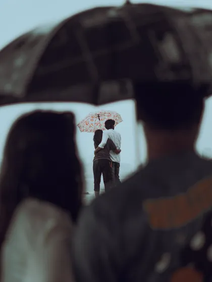 A candid, layered shot of a couple embracing under an umbrella during the Mumbai monsoon, seen through the frame of another umbrella in the foreground. It's a romantic, cinematic moment typical of the city's rainy season.