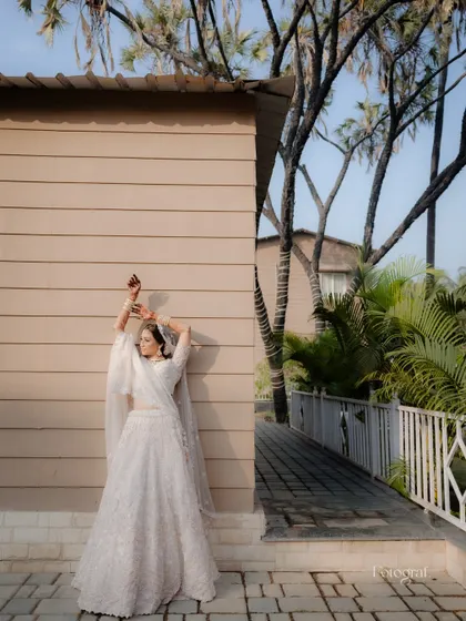A full-length bridal portrait against a modern architectural background. The bride strikes a graceful pose, showcasing her elegant white lehenga.