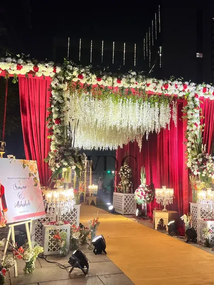A grand and dramatic entrance for a wedding. The walkway is lined with floral arrangements and candelabras, leading through an archway of red drapes and cascading white flowers.