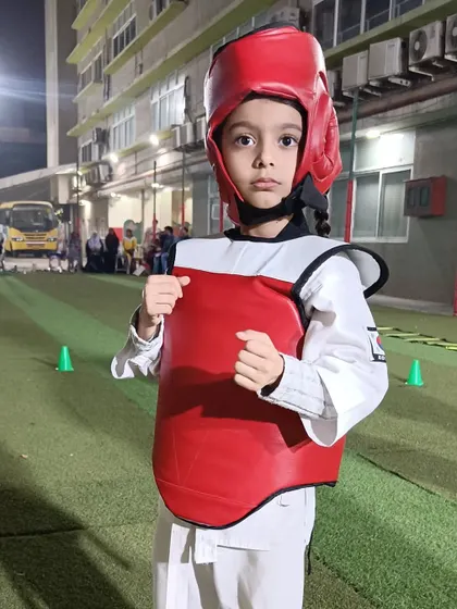 A young fighter stands ready in his full sparring gear, including the red head guard and chest protector. He is prepared for a competitive youth martial arts match.