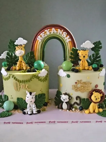 A bespoke jungle theme twin cake set for a first birthday. This design features adorable, handcrafted safari animals like a giraffe, tiger, and lion, all united by a beautiful rainbow topper.