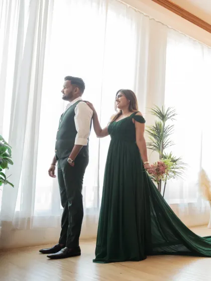 A beautifully composed shot by a large window. The bride, in her elegant green gown, places a hand on the groom's shoulder as they both look out, creating a reflective and serene mood.