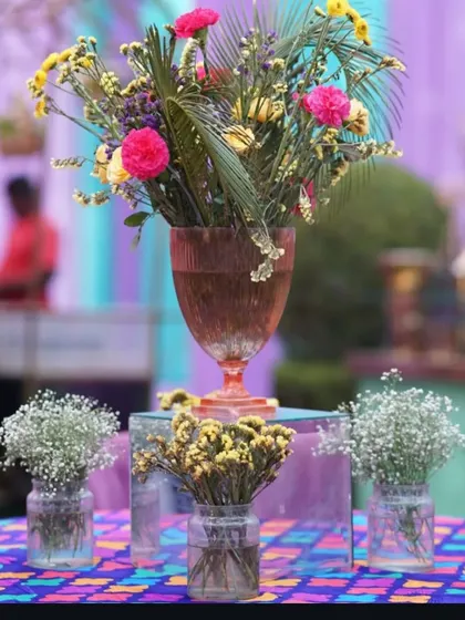 A vibrant and eclectic centerpiece arrangement. A large glass goblet holds a tropical mix of flowers, surrounded by smaller vases of baby's breath, creating a multi-level display.