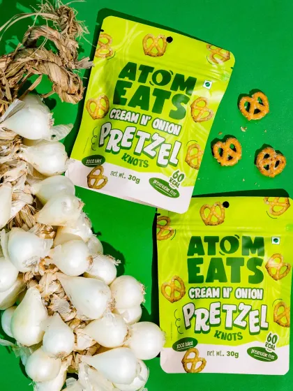 A vibrant shot of Atom Eats Cream 'n' Onion pretzels with a braid of fresh pearl onions against a bright green background. This composition creatively links the product to its core ingredient.
