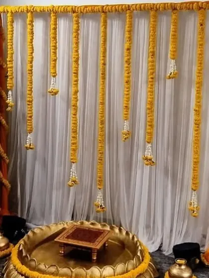 A simple and elegant Haldi setup for a home ceremony. The backdrop consists of a white curtain with hanging strings of marigold and jasmine flowers.