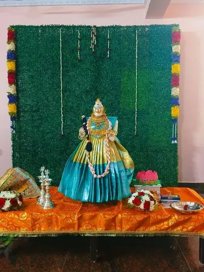 A simple Varamahalakshmi setup with a green grass backdrop and a decorated idol on an orange cloth.