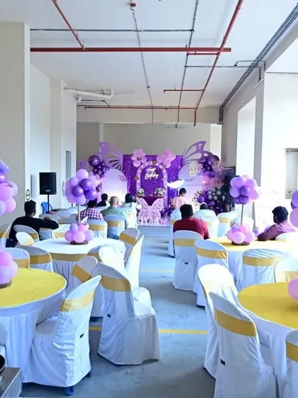 A full hall decoration for a butterfly-themed birthday party. The setup includes a main stage with a large backdrop, balloon centerpieces for each table, and coordinated chair covers to ensure a cohesive look throughout the venue.