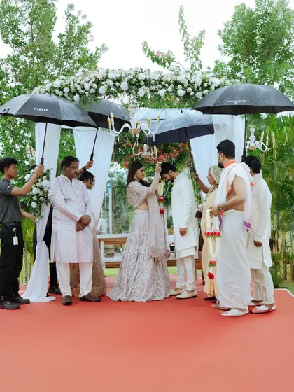 A beautiful moment during a rainy outdoor wedding, with umbrellas held over the couple as they exchange garlands.