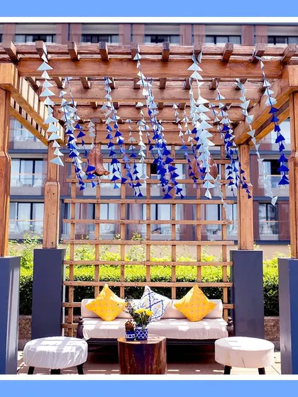 A cozy lounge seating area under a wooden pergola, decorated with hanging blue and yellow tassels. The yellow cushions add a bright, comfortable touch.