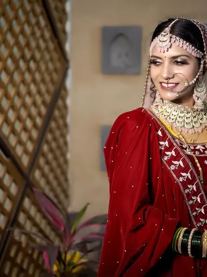 A classic bridal portrait. The bride looks away with a gentle smile, showcasing her intricate jewelry and the rich velvet of her red wedding outfit.