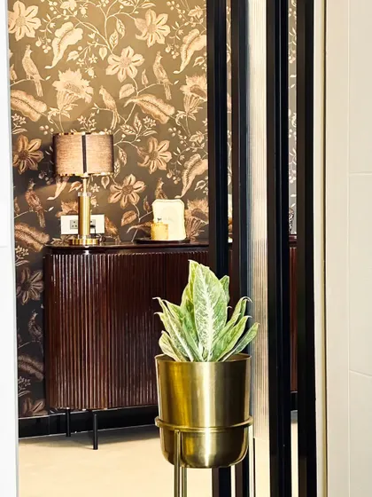 This foyer features a fluted wood console against a wall with floral wallpaper. A potted plant in a brass planter acts as a subtle divider, adding a touch of green to the warm, textured space.
