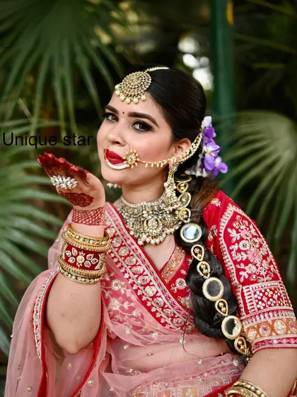 A happy bride blowing a kiss to the camera. She is wearing a traditional red and pink lehenga with a heavy gold-plated jewellery set, including a large maang tikka and a decorated braid accessory.