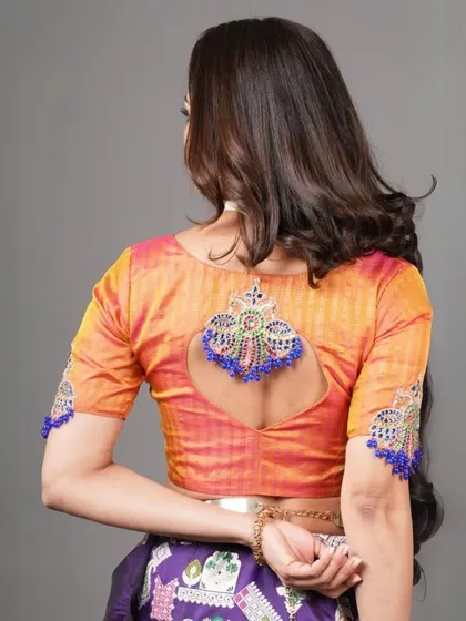 This orange and pink dual-tone blouse features a keyhole back with a beautiful tassel detail. The sleeves are also embellished, adding to its festive charm.