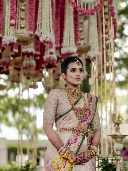 A beautiful bride standing before a traditional mandap decorated with pink and white floral tassels. The hanging brass urlis and rose drops add a touch of classic elegance to the South Indian ceremony.