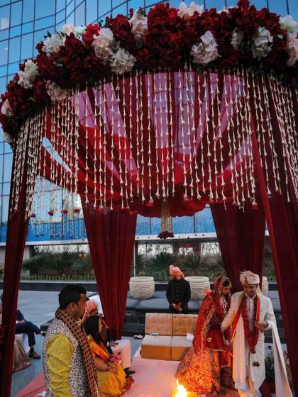 A classic round mandap design for a traditional Hindu wedding. The structure is draped in rich red fabric, with a canopy of red and white flowers and hanging tuberose strings.