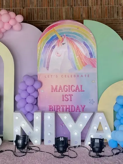 A magical first birthday setup with a unicorn and rainbow theme. The decor features pastel balloon arches, a large unicorn backdrop, and 'NIYA' in glowing marquee letters.