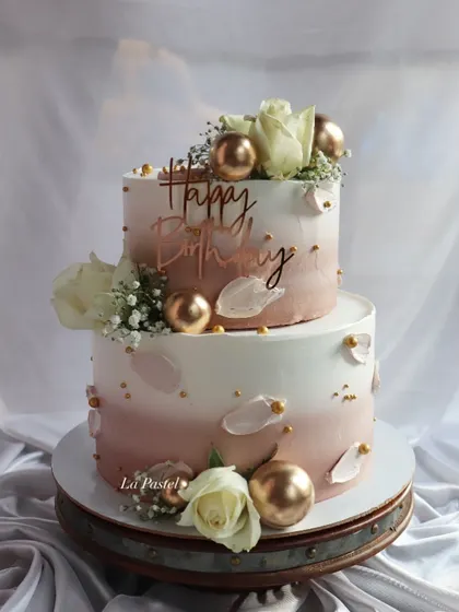 A beautiful two-tier birthday or anniversary cake with a soft pink ombre effect. It's decorated with white roses, gold spheres, and delicate baby's breath for a touch of elegance.