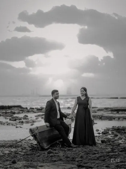A dramatic black and white photo of Akshay and Kiran on a rocky beach. The cloudy sky and the rustic boat create a moody and cinematic pre-wedding portrait.