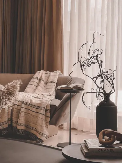 A cozy corner concept, perfect for reading. The design uses layers of texture, from the soft throw blanket to the dried botanicals and dark, sculptural vase, creating a warm and inviting nook.