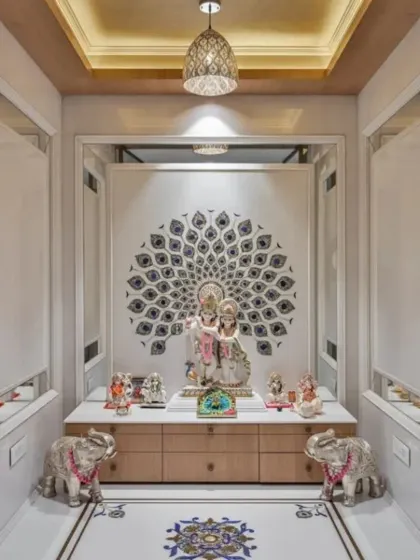 This elegant pooja room combines classic and contemporary elements, featuring a detailed peacock feather inlay on a marble wall. The space is symmetrically designed with silver elephant statues, a modern pendant light, and ample storage in the base cabinet.
