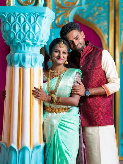 A smiling portrait of a couple in traditional attire, posing against a brightly colored, ornate pillar.