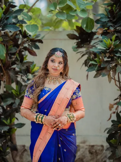 A classic bridal portrait, with the bride in a royal blue saree, looking elegant against a natural green backdrop.