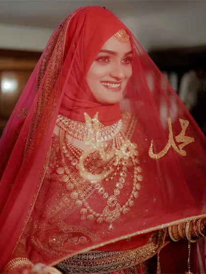 A stunning close-up of a Muslim bride, her face illuminated with joy, seen through her traditional red veil adorned with sacred calligraphy.