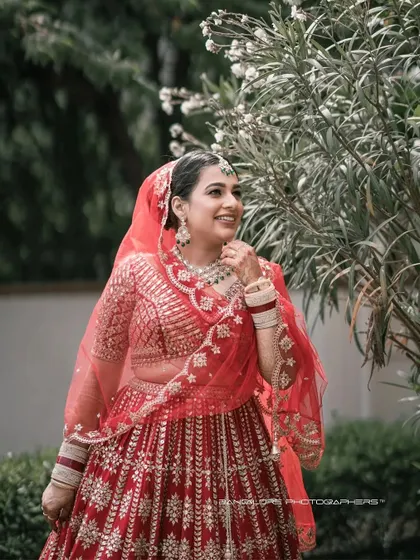 A candid shot of the bride smiling, her red lehenga vibrant against the soft green background. This is a moment of pure, unposed happiness.