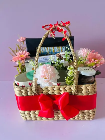 A beautiful couple's hamper in a cane basket. It includes heart-themed mugs, snacks, and floral decorations, perfect for an anniversary or wedding.