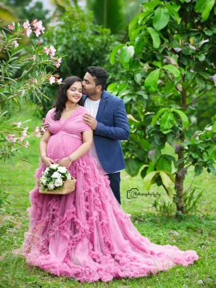 A sweet couple's portrait in a garden, with the father-to-be kissing the mother-to-be's forehead. She is wearing a pink ruffled gown.