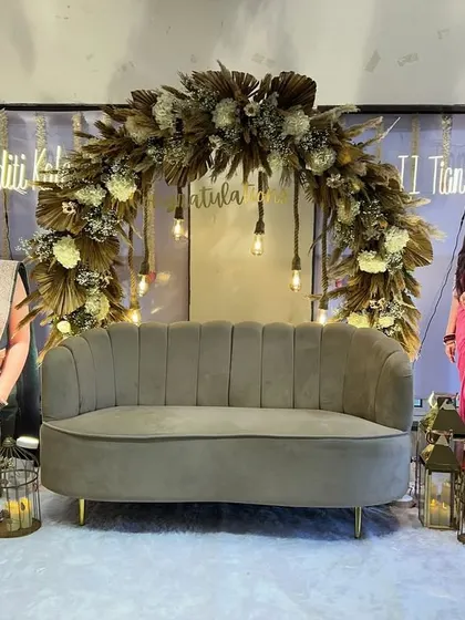 A unique boho-chic setup to celebrate a major milestone for two sisters. The dried floral arch, hanging Edison bulbs, and plush velvet sofa create a sophisticated and celebratory stage.