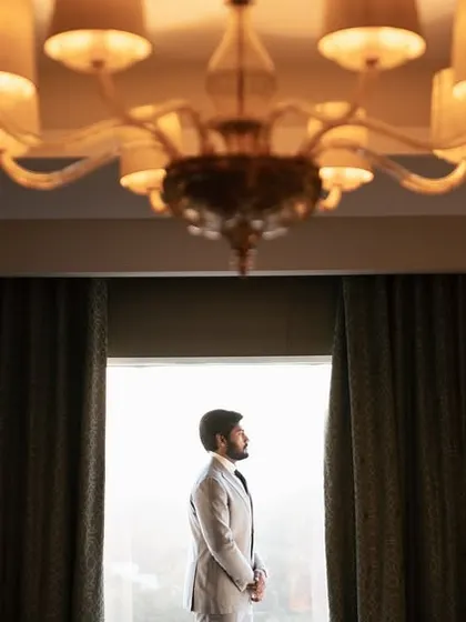 A cinematic shot of the groom standing in a hotel room, silhouetted against a large window, creating a dramatic and stylish portrait.