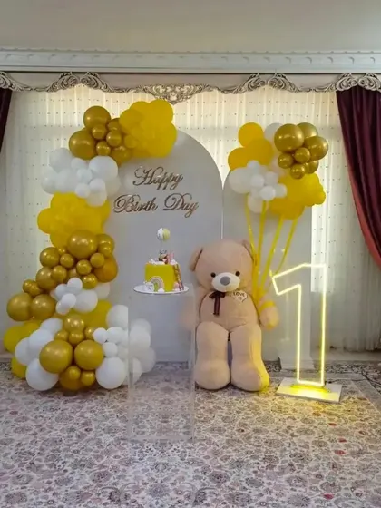 An elegant first birthday setup in yellow and white. This home decoration features a balloon arch against a custom backdrop, a large teddy bear, and a glowing number '1'.
